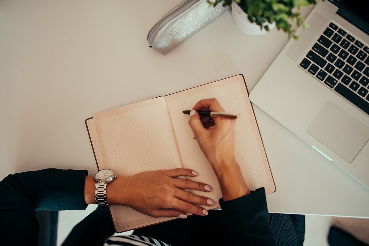 A person writing in a notebook beside an open laptop. Editorial photograph, not a Quilty score.